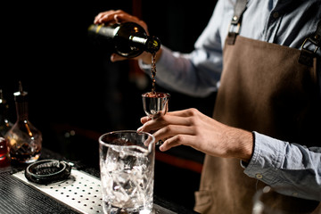 Professional male bartender pouring a brown alcoholic drink from the bottle to a steel jigger