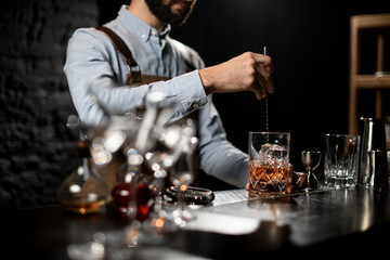Professional bartender stirring a brown cocktail with ice cubes in the glass with special spoon