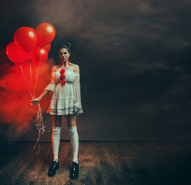 portrait cute young woman with scary face clown grimm in white old dress stands in backdrop dark black gothic room holding red balloons. art creative makeup. Fog smoke free space text. lady ghost