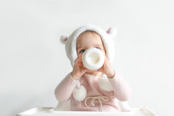Little toddler girl in a warm fluffy hat drinks milk from a bottle while sitting. Half-length...