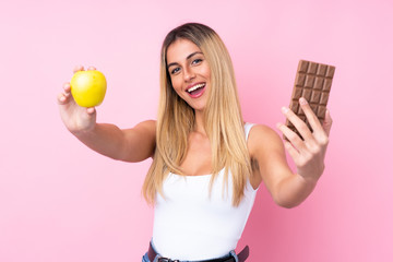 Obraz premium Young Uruguayan woman over isolated pink background taking a chocolate tablet in one hand and an apple in the other