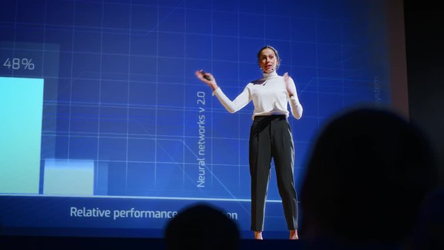 On Stage, Successful Female Speaker Presents Technological Product, Uses Remote Control for Presentation, Showing Infographics, Statistics Animation on Screen. Live Event / Device Release. Slow Motion