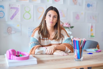 Young beautiful teacher woman wearing sweater and glasses sitting on desk at kindergarten with...