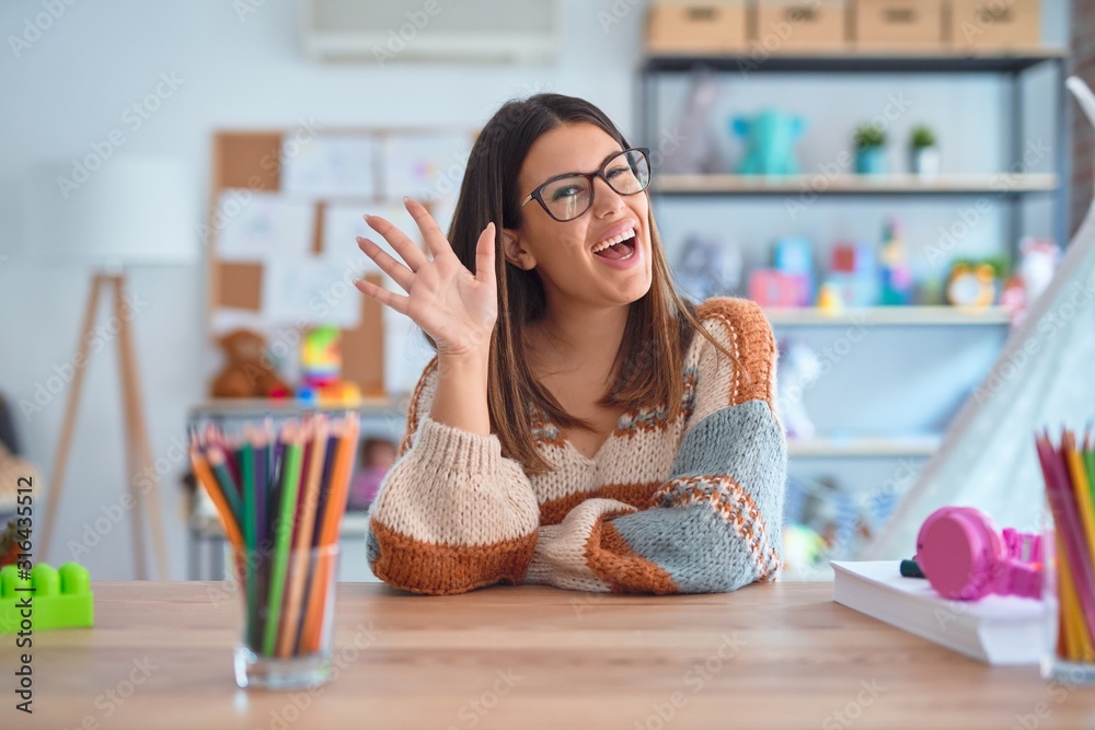 Wall mural young beautiful teacher woman wearing sweater and glasses sitting on desk at kindergarten waiving sa