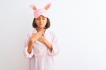 Beautiful child girl wearing sleep mask and pajama standing over isolated white background smiling with hands on chest with closed eyes and grateful gesture on face. Health concept.