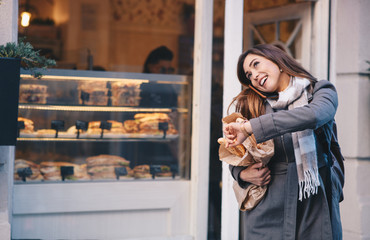 A young woman in front of a bakery holding bag of pastry products, talking on the phone.