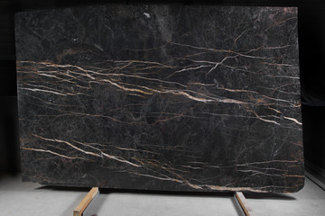 A slab of brown marble with light veins stands on a rack, called Port Laurent