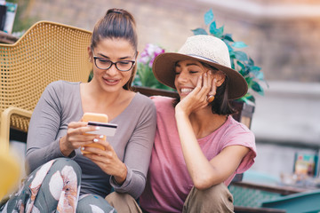 Two women sitting outdoors, shopping online via phone.