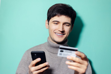 Studio portrait of young happy man with smartphone and credit card in hand on background of aqua menthe color.