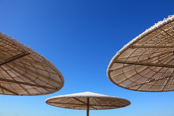 Fototapeta premium Tropical beach sun shades against bright blue sky on a sunny day. Up view. Sun umbrellas of reed and straw. Travel and vacation. Background with copy space