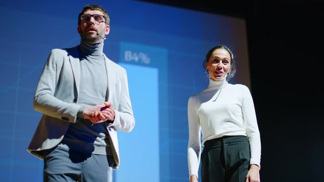 On Stage, Successful Female Executive and Male Manager Present New Product, Show Infographics, Statistics on Big Screen, Talk About Growth. Live Event, Tech Startup, Business Conference