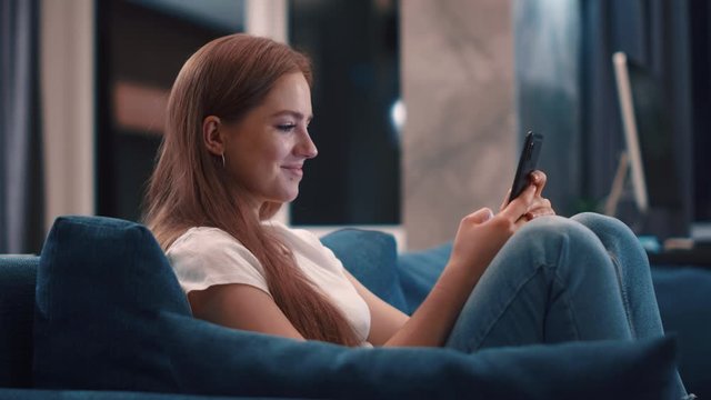 Relaxing cheerful young woman with golden hair sitting on blue sofa uses phone smile at modern apartment texting share messages applications on social media enjoy feel happy mobile technology evening