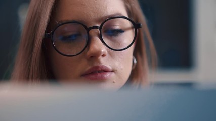 Close up view of attractive young redhair woman with glasses working on project by laptop computer managing distant job duties staying at home in comfy modern interior at evening. Freelance concept. - Powered by Adobe