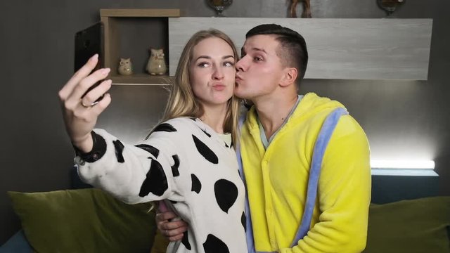 Young funny couple in pajamas taking selfie photos with smartphone camera standing in living room. Concept of happiness and togetherness. Medium shot in slow motion