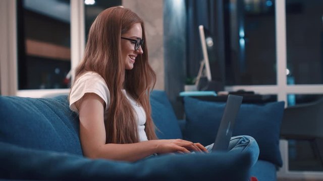 Side view of happy red hair young woman in white t-shirt sitting on blue coach browsing internet chatting with friends on modern laptop smile resting at modern apartment having fun evening relax
