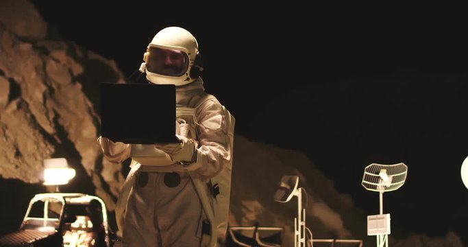 Caucasian male astronaut using laptop computer for explorations on the Mars at night or having videochat with family from space. Outdoor.
