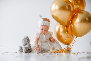 A one year old, baby girl sitting with a bunch of golden balloons and taddy bear on white...