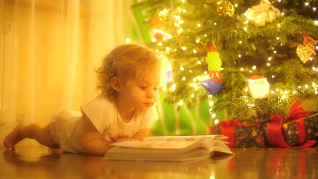Happy blonde baby girl looks at pictures in a fairytale book near decorated Christmas tree