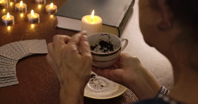 Shooting over shoulder, mature Caucasian woman holding cup with coffee sediment and talking. Mature lady reading fate on coffee grounds. Tassology, fortune telling.. Cinema 4k ProRes HQ.