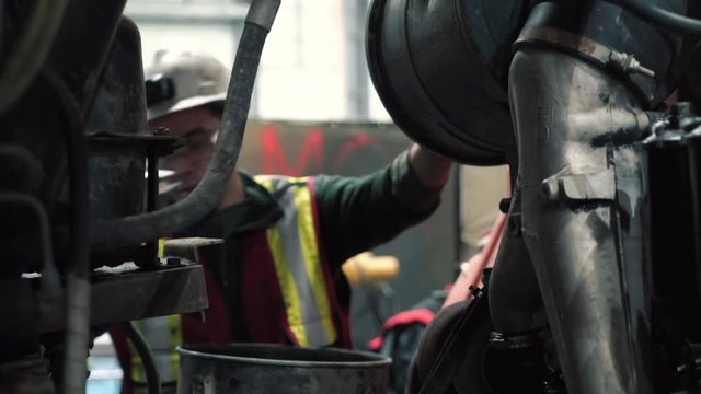 The mechanic changes the filter in the truck. He wears a helmet, a reflective vest, glasses and gloves. It's for safety.