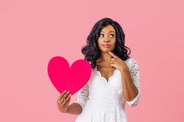 Portrait of  young woman with black curly hair holding pink heart looking up and thinking,...