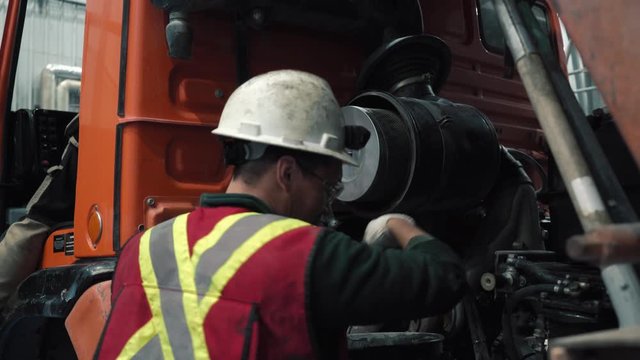 The mechanic changes the filter in the truck. He wears a helmet, a reflective vest, glasses and gloves. It's for safety.