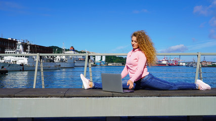 Smiling blonde works at a computer on a bench near the ocean. Freelancer on an observation deck against the background of the sea and clear sky. Sunny day taiwan