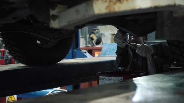 The mechanic repairs the car.  He picked it up on a jack. He's gonna change the oil, and he's examining it. He's wearing glasses, a reflective vest and a helmet. It's for safety.