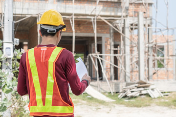 Back view of Thai young male engineer or architect wearing yellow-orange vest, yellow and safety helmet holding and looking at construction paper plan in front of a new building unfinished house.