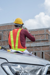 Thai young male engineer or architect wearing yellow-orange vest and yellow safety helmet is sitting on a car and talking to phone in front of unfinished house.