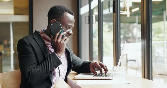 Young African Businessman talking on the mobile phone with business clients in office using laptop by the window freelance Black man checking information on computer chatting on phone in cafe
