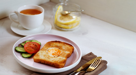 Brunch - an egg with sesame seeds on toast, fresh cucumber and tomato. A cup of tea with lemon. In the background is a can of sliced lemon with sugar. Cutlery lie on a textile napkin.