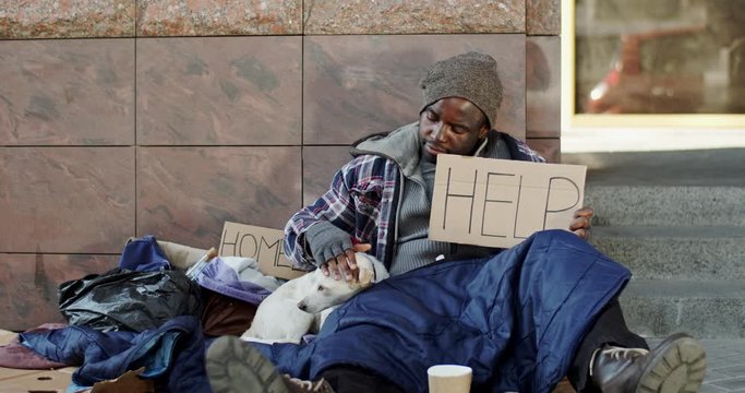 Portrait Shot Of The Young Poor African American Man Beggar Sitting On The Ground In The Urban Area With A Table Help And Homeless. Outside.