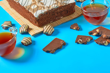 One whole checkered rectangular cake near broken chocolate bar, candy and two full glass cups of tea lies on blue table on party. Celebration concept. Space for text