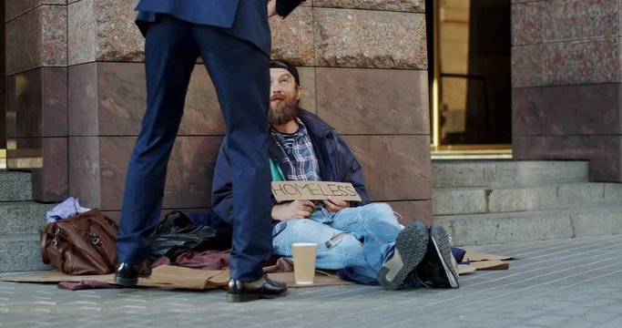 Young Caucasian homeless poor and dirty man sitting on the street and asking for money when businessman coming to him, giving food and talking to the man. Outside.