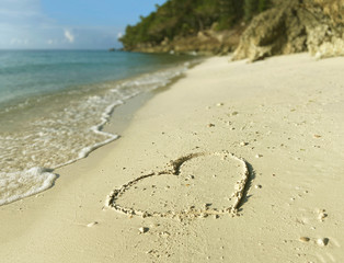 Closeup heart drawn on a sandy yellow beach. White sea foam against the background of the sea, blue sky and rocky shore with trees.
