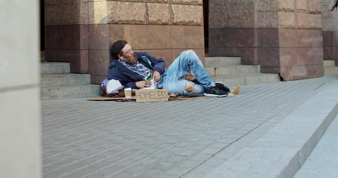 Poor and dirty Caucasian man, homeless alcoholic lying on the ground at the street in the city while successful people walking by.
