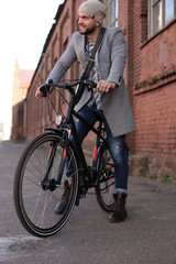 Handsome young man in grey coat and hat standing with his bicycle.