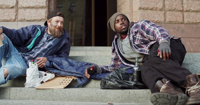 Two mixed-races poor and dirty homeless men lying on the ground of the street and talking while collecting money on food.