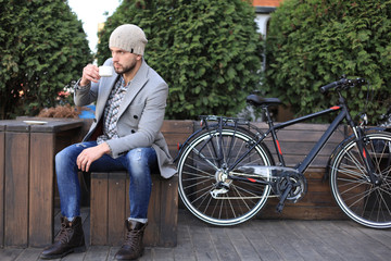 Handsome young man in grey coat and hat sitting on a bench relaxed drinking coffee and thinking near his bicycle.