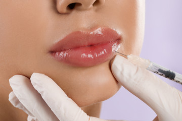Young woman getting lips injection on lilac background, closeup