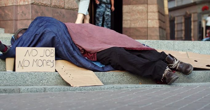 Portrait of the young African American male homeless beggar sleeping on the ground with a cup for money and table No Job No Money.