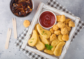 Buttered chicken nuggets and popcorn bites in white vintage wooden box with ketchup and glass of cola on light background. Top view
