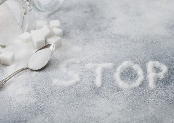 Glass jar of natural white refined sugar with cubes with silver spoon on light table background with STOp letters. Unhealthy food concept.