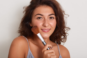 Close up portrait of energetic positive delighted female looking directly at camera, holding makeup brush for blush in one hand, applying product to cheeck, isolated over white background in studio.