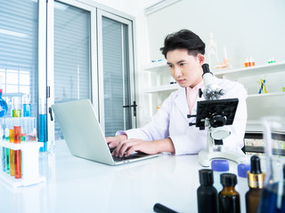 Obraz na płótnie Canvas Asian young man scientist concentrate in research and test biology or chemistry innovation with computer laptop technology device for analysis discovery information in clean laboratory room