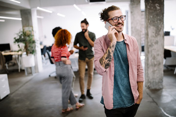 Happy young man looking at his mobile phone and smiling while his colleagues working in office