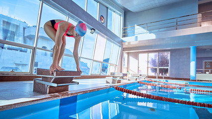 Sporty athletic female champion swimmer in low position on starting block in a swimming pool during the sports competition
