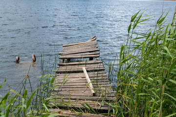 Fototapeta premium Remnants of the old abandoned wooden bridge. Desolate pier. Forest and sky reflection on water. Shore of a single lake