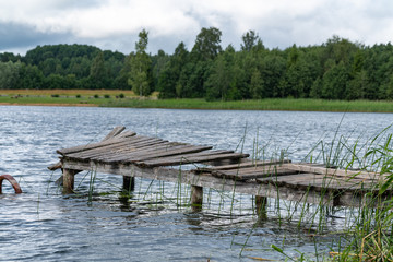 Remnants of the old abandoned wooden bridge. Desolate pier. Forest and sky reflection on water. Shore of a single lake